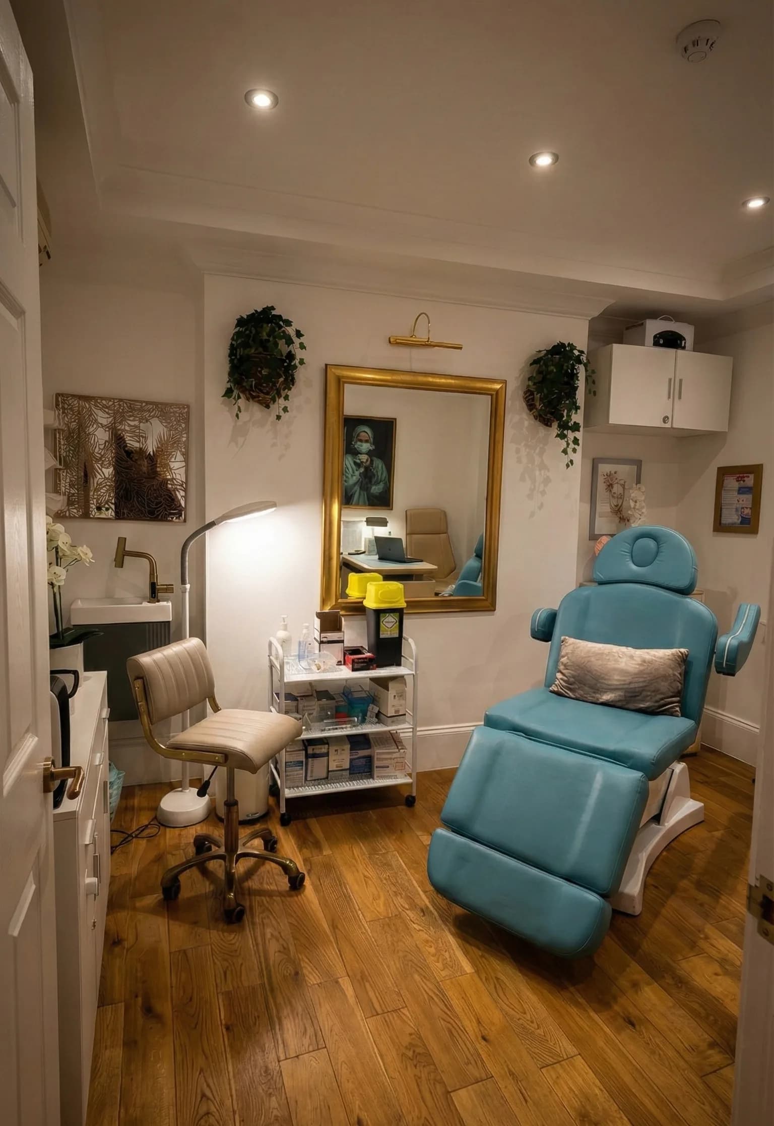 A treatment room at the LV8 London clinic: a blue reclining treatment chair with a cushion, a gold-framed mirror, a stocked supplies trolley and an oak floor