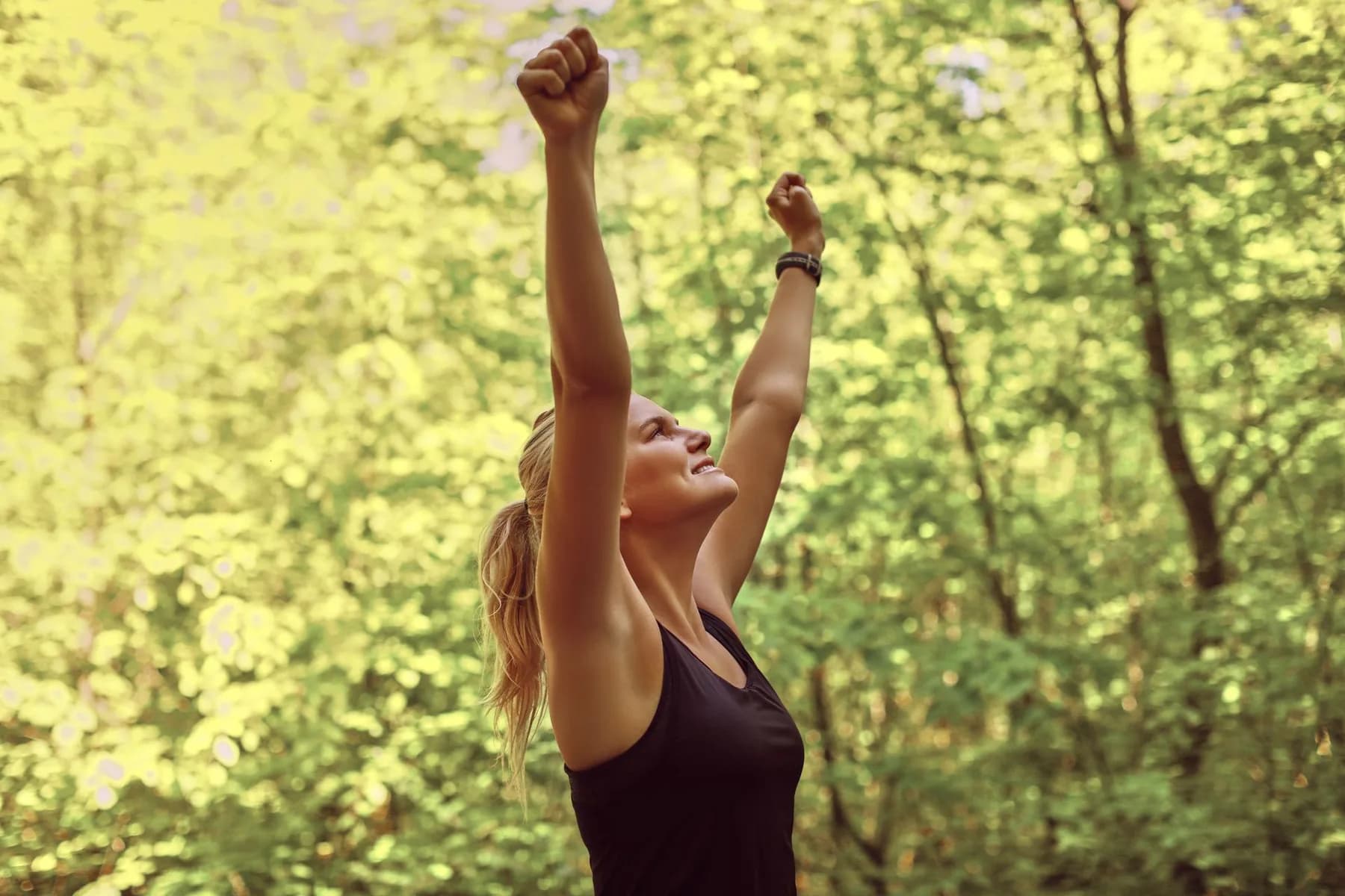 A woman in sunlit woodland with both arms raised above her head, wearing a black sports watch