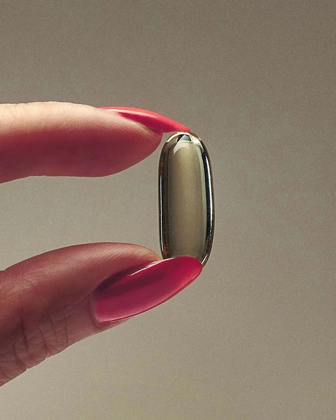 A single polished gold capsule held between two fingertips against a warm neutral backdrop