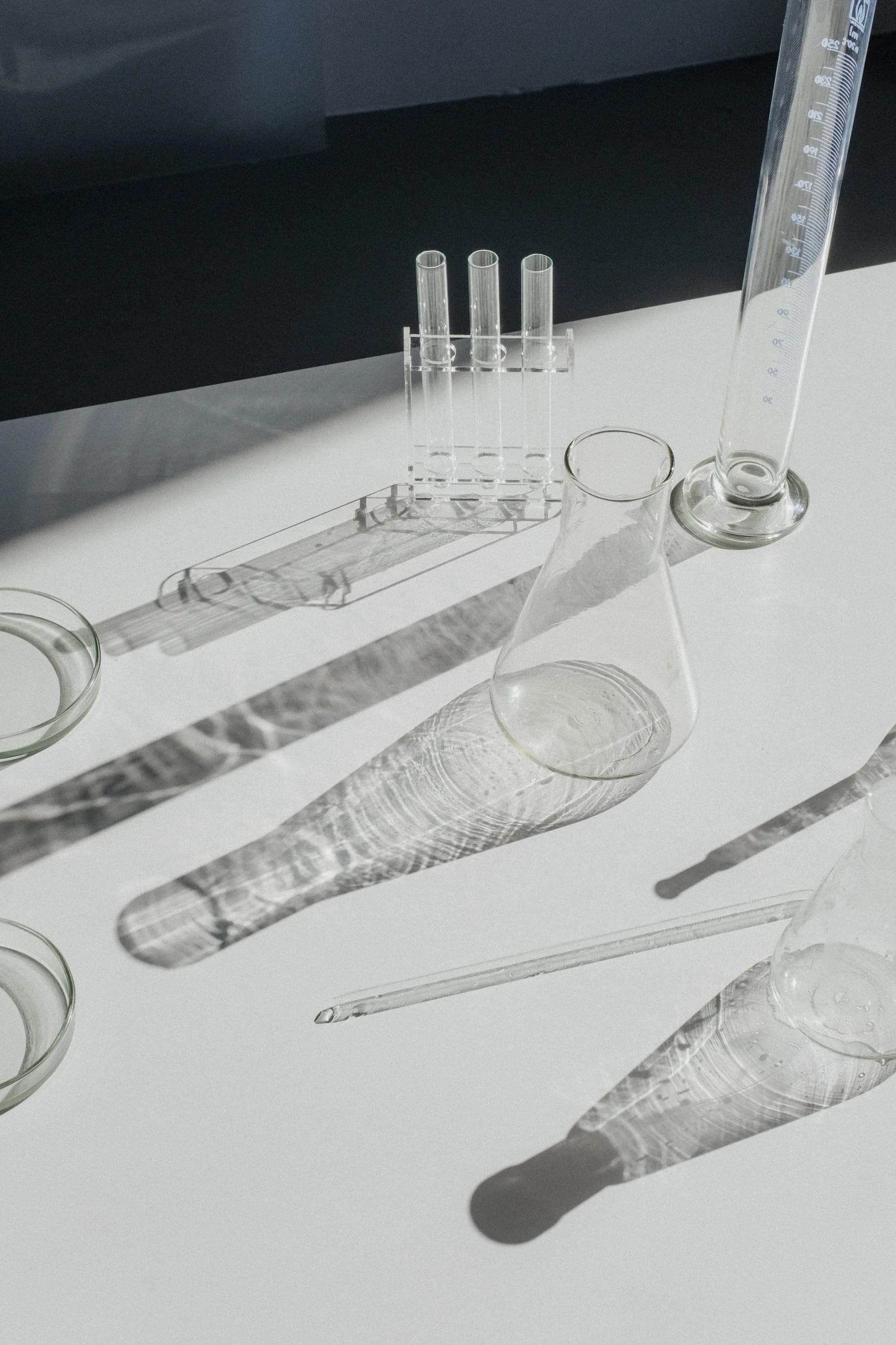 Laboratory glassware, test tubes, a graduated cylinder and flasks on a white bench, casting long shadows in hard daylight