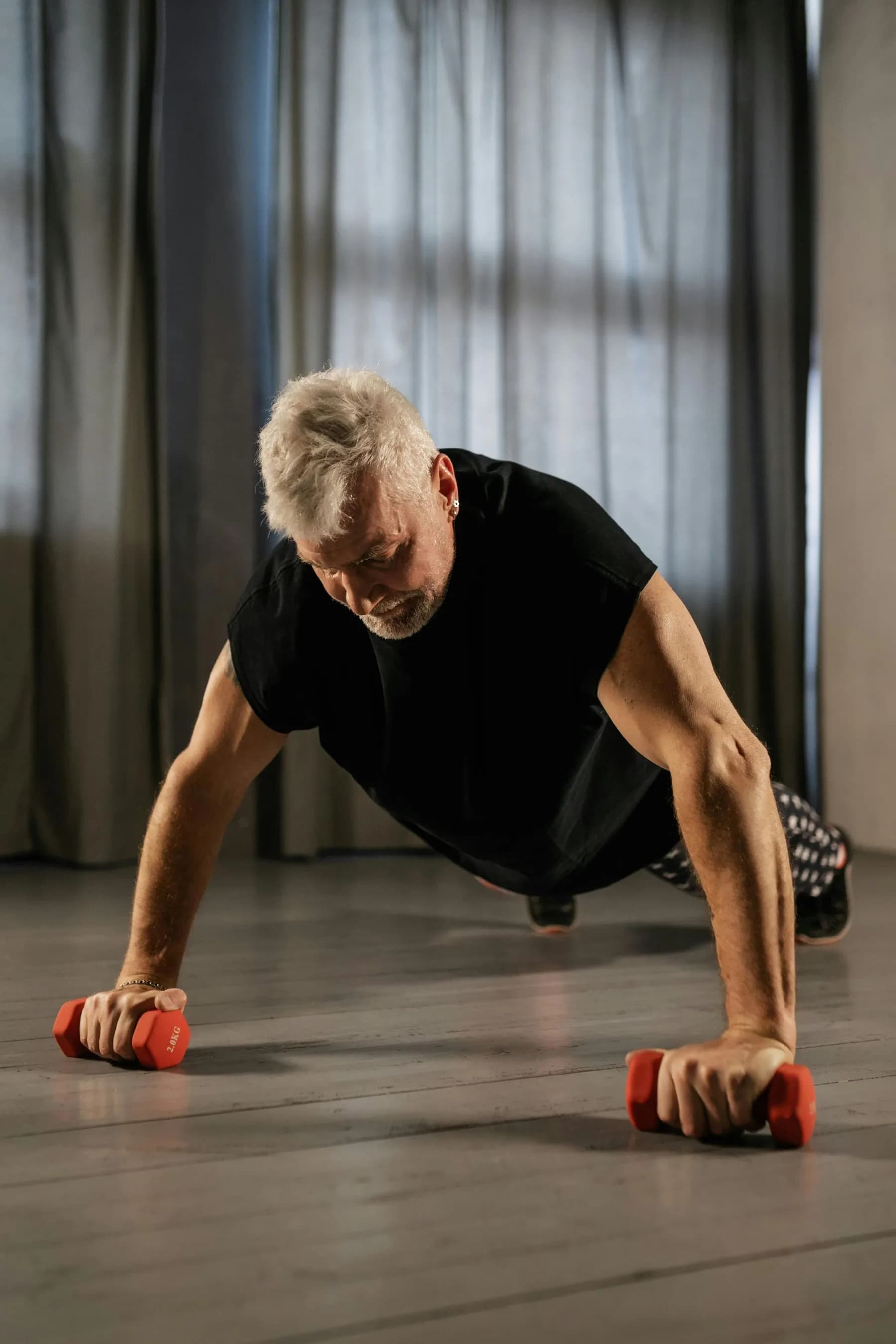 A silver-haired man in a black t-shirt holding a press-up on a pair of red dumbbells in a curtained studio