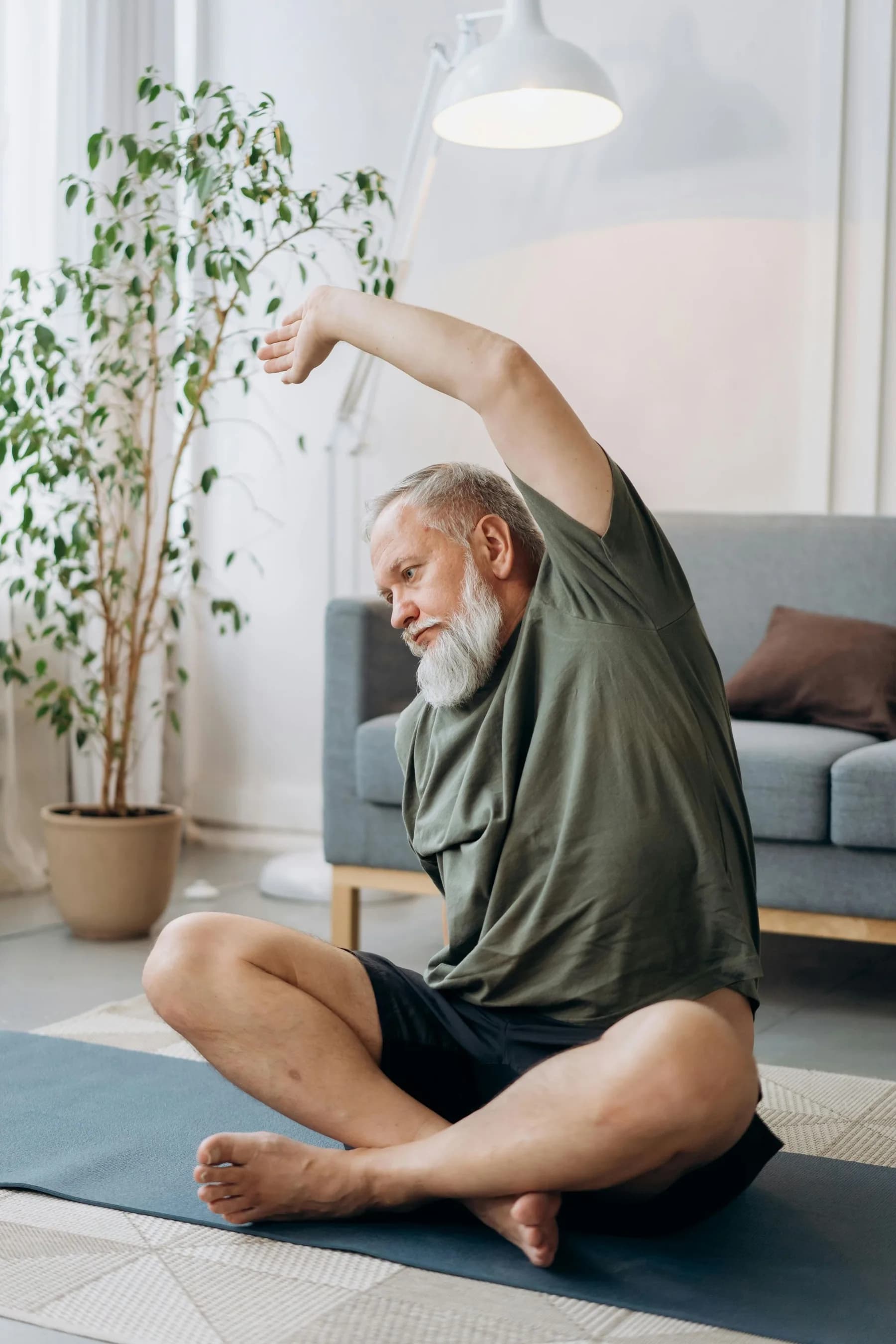 An older man sitting cross-legged on a yoga mat in a bright living room, stretching one arm overhead