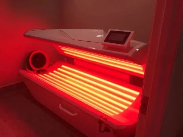 An empty white red light therapy pod with its canopy raised, horizontal lamp banks glowing warm red across the bed inside a small treatment room
