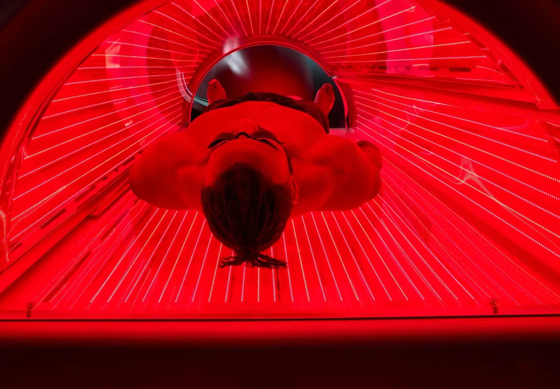 Overhead view of a person lying inside a full-body red light bed, the LED strips above and below flooding the enclosure with deep red light